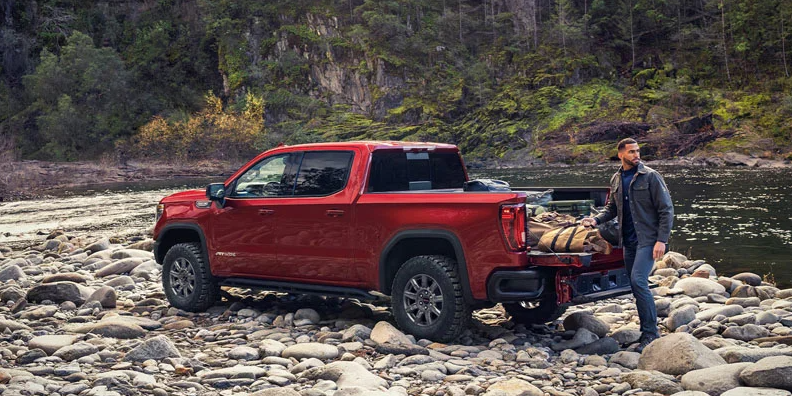 A red 2026 GMC Sierra 1500 parked on a river bank with a man unloading camping gear from the back.